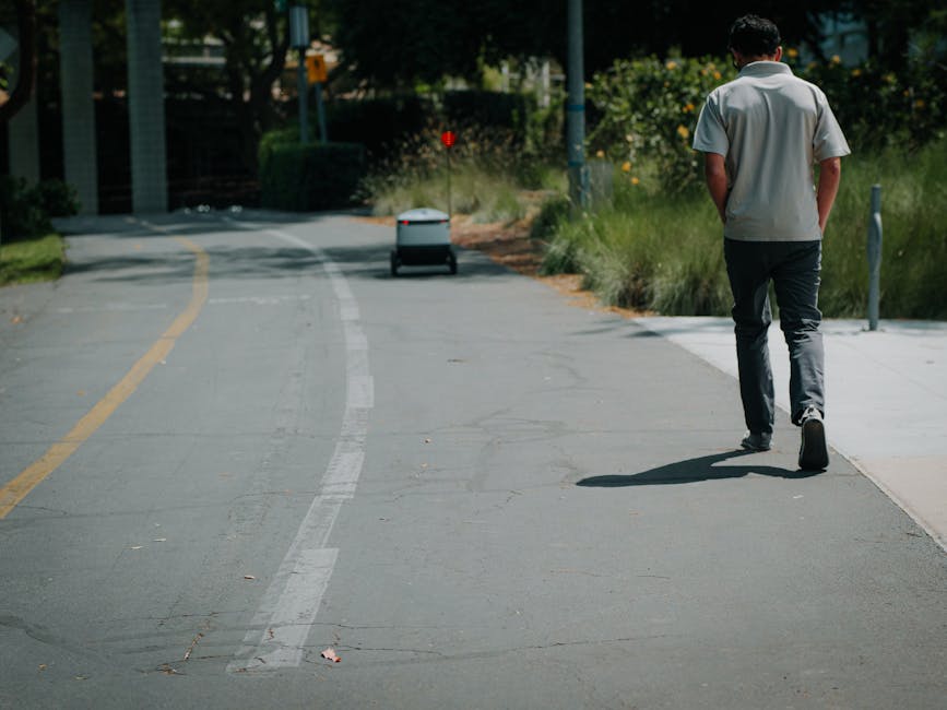 A young man walking along a paved pathway with a slight bend, wearing a light grey short-sleeved shirt and dark trousers, seen from behind. On the left side of the image, a white delivery robot with r