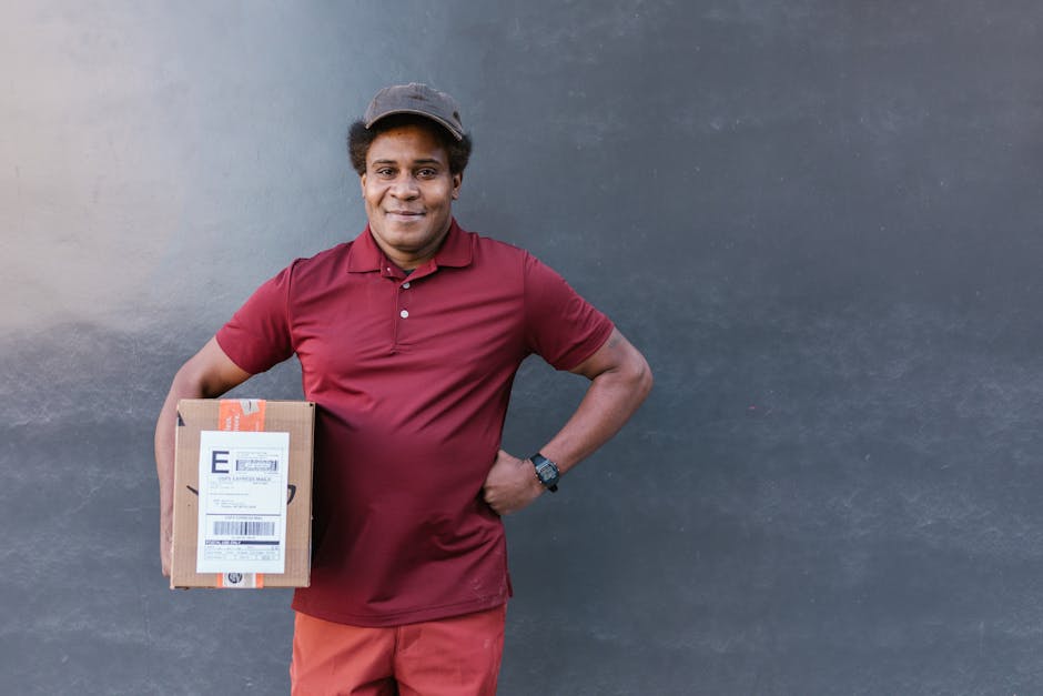 A smiling delivery person stands against a plain dark grey background, holding a small cardboard parcel with a shipping label in his left hand. He is dressed in a maroon polo shirt and matching trouse