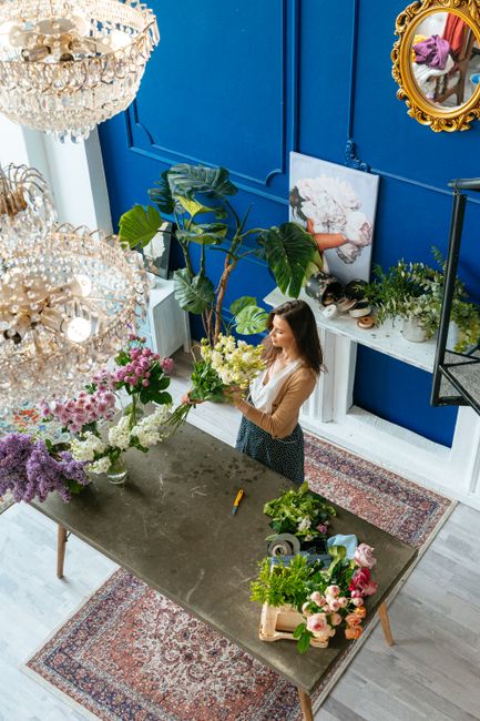 A woman arranging a bouquet of fresh flowers inside a cozy floral shop with vibrant blue walls, decorated with a large crystal chandelier hanging from the ceiling. The bouquet features a variety of bl