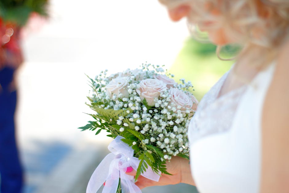 A person holding a delicate floral bouquet featuring pale pink roses and white baby's breath, arranged with lush green fern leaves. The bouquet is wrapped with a white ribbon and partly covered with t